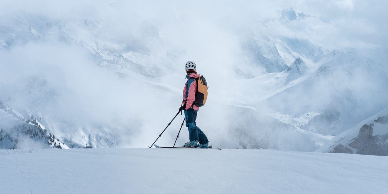 man poses in his skiing attire with background of snowy mountains